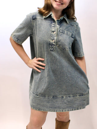 Woman wearing a washed denim short sleeve collared mini dress with front buttons and chest pocket.