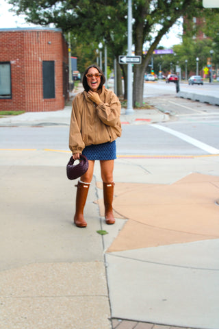 A woman stands on a city sidewalk wearing a tan, oversized, bubble hem jacket with a blue patterned mini skirt, brown rain boots, and holding a dark purple handbag. The background shows a street, trees, and a brick building.