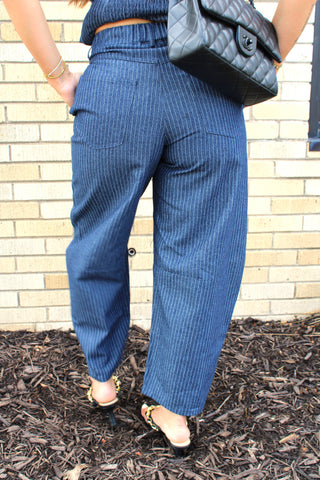 A person shown from the back wearing blue pinstriped wide-leg barrel trousers, and carrying a black quilted shoulder bag.