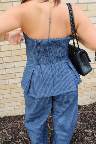A woman shown from behind wearing a blue pinstriped smocked strapless top and matching pants, carrying a black quilted shoulder bag against a beige brick wall.