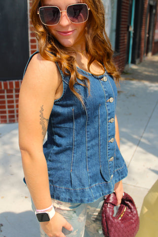 A woman wearing a sleeveless blue denim vest with silver buttons, paired with distressed jeans and sunglasses, holding a burgundy quilted handbag on a city sidewalk.