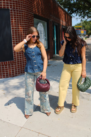 Two women walking on a sunny sidewalk next to a red brick building. The woman on the left wears a denim vest over a light blue floral flared jeans, holding a burgundy textured handbag.