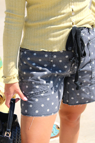 Close-up of a person wearing dark black washed denim shorts with white polka dots and a black braided drawstring waist.