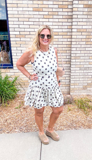 Woman wearing a black and white polka dot sleeveless mini dress with a drop waist and flared skirt, styled with sunglasses and wedge sandals