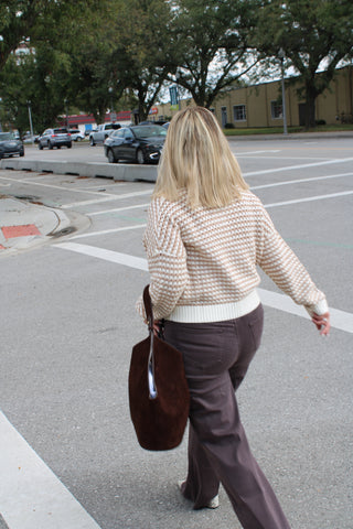 A woman walking away on a paved street, wearing a cream and brown patterned knit sweater, brown pants, and carrying a large dark brown tote bag.