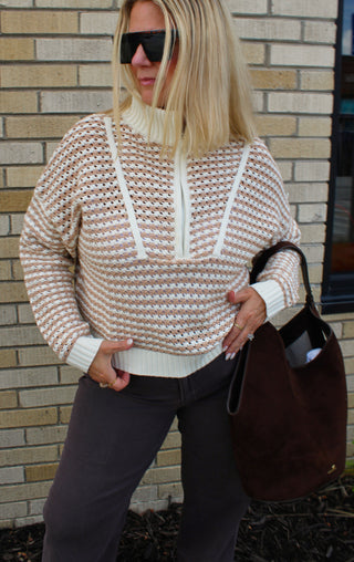 A woman wearing a beige and white patterned knit mock-neck sweater, dark brown pants, and black sunglasses, holding a brown suede tote bag against a brick wall background.