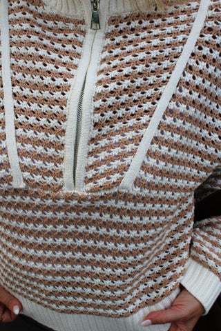 Close-up of a hand-knit half-zip sweater with a textured waffle pattern in beige and white, featuring cream-colored drawstrings and a silver zipper.