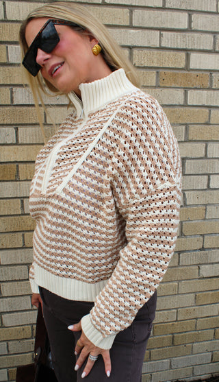 A woman wearing a cream and brown striped knit turtleneck 1/4 zip sweater with a textured openwork pattern, styled with dark pants and sunglasses, standing against a brick wall.