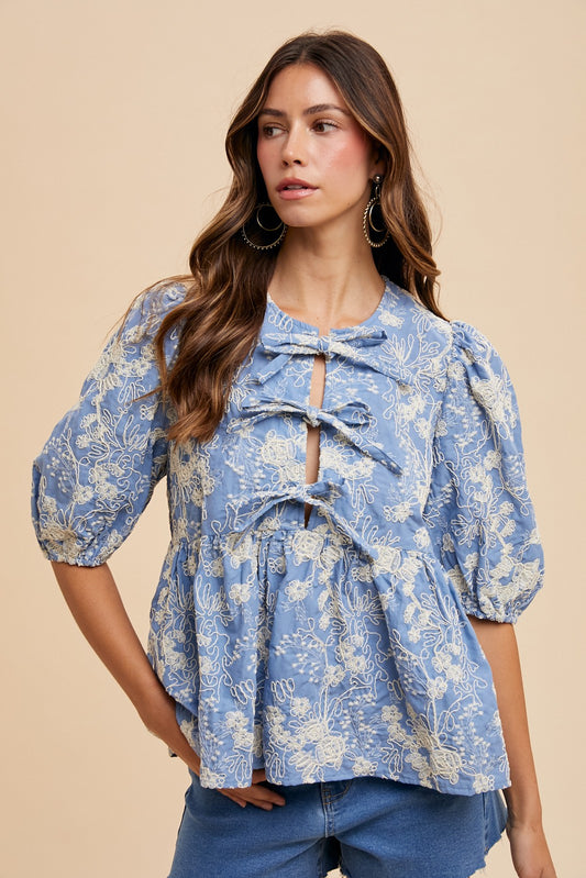 Woman wearing a light blue puff sleeve blouse with ivory floral embroidery and three bow front ties – romantic spring peplum top for women.