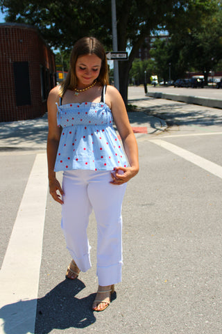 A woman standing on a city street wearing a light blue polka-dot ruffled top, white pants, and tan sandals.