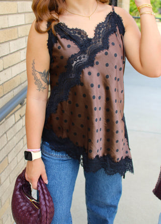 A woman wearing a brown camisole top with black polka dots and black lace trim, paired with blue jeans and holding a purple quilted clutch bag.
