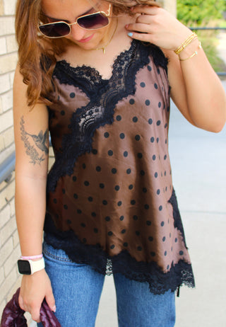 A woman wearing a sheer brown polka-dot camisole top with black lace trim, styled with blue jeans, gold-framed sunglasses, and bracelets.