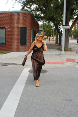 A woman walking on a city street wearing a sheer black lace camisole and matching skirt with a brown polka-dot pattern underneath, holding a small black handbag.