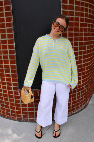 Woman wearing Camden Stripe Button Knit Sweater featuring yellow and blue stripes, relaxed fit, long sleeves, and button henley neckline styled with white pants.