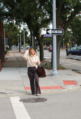 A woman walking on a city sidewalk near a street crossing, wearing a cream and khaki striped long-sleeve top, dark pants, and carrying a brown tote bag.