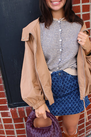 A woman wearing a Camel brown lightweight jacket with elastic cuffs over a striped button-up sweater vest, paired with a denim blue patterned skirt, holding a purple woven handbag against a brick wall background.