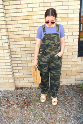 Woman wearing camouflage overalls and sunglasses, standing against a brick wall.