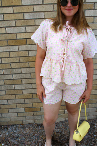 Woman wearing Carmin Floral Gingham Top featuring pink gingham print, floral embroidery, tie-front detail, flutter sleeves, and peplum silhouette.