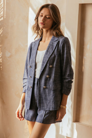 Woman wearing a chambray blazer with silver button details and matching tailored shorts, styled with a textured cream knit tank, standing in soft natural lighting.