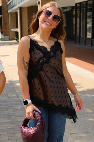 A woman wearing a black polka-dot camisole top with lace trim, blue jeans, and sunglasses, holding a woven burgundy handbag on a city sidewalk.