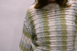 A close-up of a person wearing a chunky, open-knit crochet sweater with horizontal stripes in sage green and off-white for women available at Erin Rose Boutique.