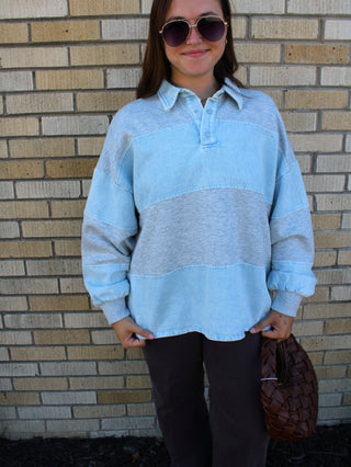 A woman wearing a light denim blue and heather gray striped long-sleeve rugby-style shirt, dark pants, and sunglasses, standing in front of a tan brick wall and holding a brown woven bag.