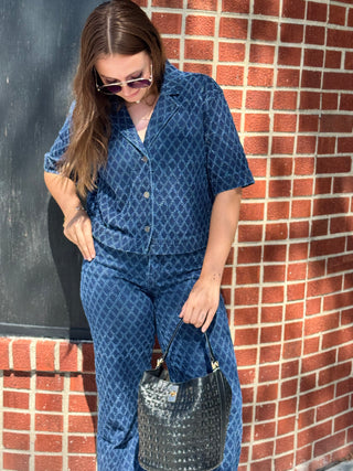 A woman wearing a blue diamond textured short-sleeve button-front top and matching pants, standing against a red brick wall and holding a black textured handbag.
