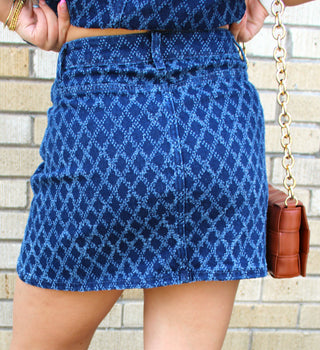 A person wearing a blue patterned denim mini skirt and matching top, holding a brown quilted handbag with a gold chain strap, standing against a light brick wall.