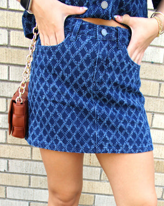 A close-up of a person wearing a blue denim mini skirt with diamond textured pattern, holding a brown leather woven handbag.