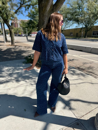 A woman stands on a sunny sidewalk wearing a matching navy blue patterned short-sleeve top and wide-leg pants set, holding a black textured handbag.