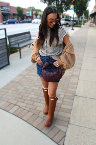 A woman stands on a city sidewalk wearing a gray striped top, a camel jacket draped off her shoulders, the dara blue diamond textured mini skirt, and brown knee-high hunter rain boots, while holding a dark purple woven handbag.