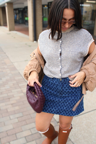 A woman wearing a blue diamond textured patterned mini skirt, a  striped top, and a tan jacket, holding a purple clutch bag while walking on a paved sidewalk in hunter camel rain boots