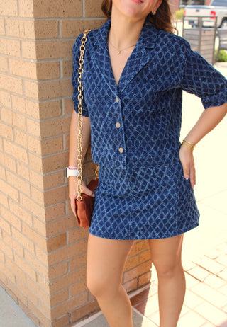 A woman wearing a blue denim diamond textured short-sleeve blazer and matching mini skirt, standing outdoors next to a tan brick pillar.