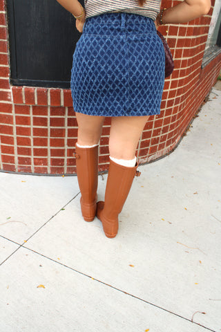 A person shown from the back wearing a blue diamond textured mini skirt, brown knee-high rain boots, and a striped top, standing on a sidewalk in front of a red brick building.