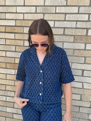 A woman wearing a blue diamond patterned short-sleeve button-up denim shirt and matching pants & wearing dark sunglasses.