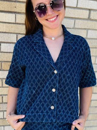 A woman wearing a dark blue short-sleeve button-front shirt with a small white geometric pattern, standing against a light brick wall.