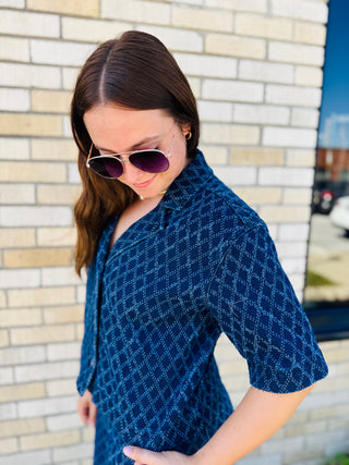 A woman wearing a blue diamond patterned short-sleeve button-up denim shirt and aviator sunglasses, standing in front of a light-colored brick wall.