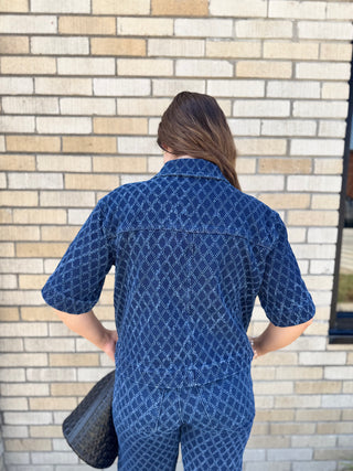 A woman viewed from behind wearing the dara dark blue diamond textured short-sleeve shirt and matching pants, standing in front of a beige brick wall and holding a black bag.