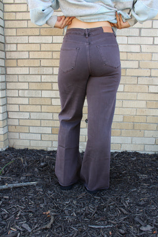 A person viewed from behind wearing loose, wide-leg Mocha Brown Dear John Fiona jeans, standing outdoors against a light brick wall with mulch on the ground.