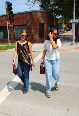 Two women walking across a street crosswalk outdoors. The woman on the left wears a metallic bronze-brown satin sleeveless top with black lace trim and blue jeans, holding a dark burgundy clutch. Woman on right wearing dear john taylor starboard denim jeans and lace top.