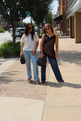 Two women walking on a sunny outdoor sidewalk next to a street with trees and storefronts. The woman on the left wears a white patterned short-sleeve top, light blue jeans, and brown sandals, holding a dark purple handbag.
