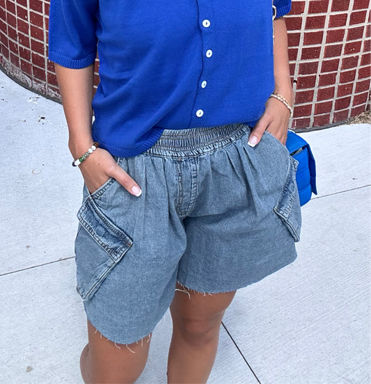 Woman wearing washed blue denim cargo shorts with an elastic waistband, oversized pockets, and frayed hem, styled with a cobalt blue top