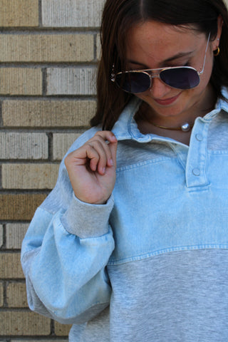 A woman wearing a denim light blue and heather gray color-blocked pullover sweatshirt with denim collar, styled with aviator sunglasses, standing against a beige brick wall.