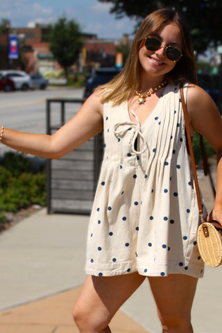 Model wearing the Dot & Dandy Polka Dot Denim Romper featuring a cream denim fabric, navy polka dots, pintuck detailing, tie-front neckline, and relaxed babydoll silhouette.