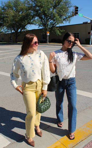 Two women walking on a sunny city street crosswalk. The woman on the left wears a cream high-neck long-sleeve top with blue floral embroidery, light yellow pants, and a green woven handbag.