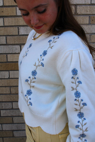 A woman wearing a white knit sweater with blue floral embroidery down the front and sleeves.