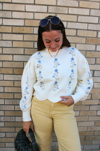 A woman wearing the Flora Embroidered Knit Sweater in ivory with blue floral embroidery down the front and on the sleeves, paired with yellow pants.