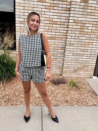 Woman wearing a black and white gingham matching set with a sleeveless boxy top and high-waisted shorts, styled with black heels and a structured handbag.

