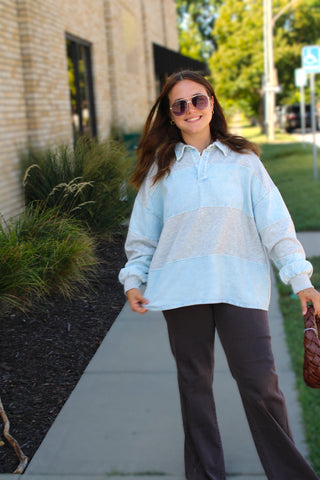 A woman wearing a light blue and heather gray color-blocked long-sleeve polo shirt and dark brown pants, standing on a sidewalk outdoors.