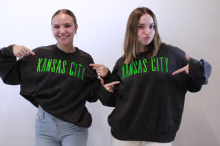 Two people wearing black sweatshirts with 'Kansas City' printed in green on a white background.